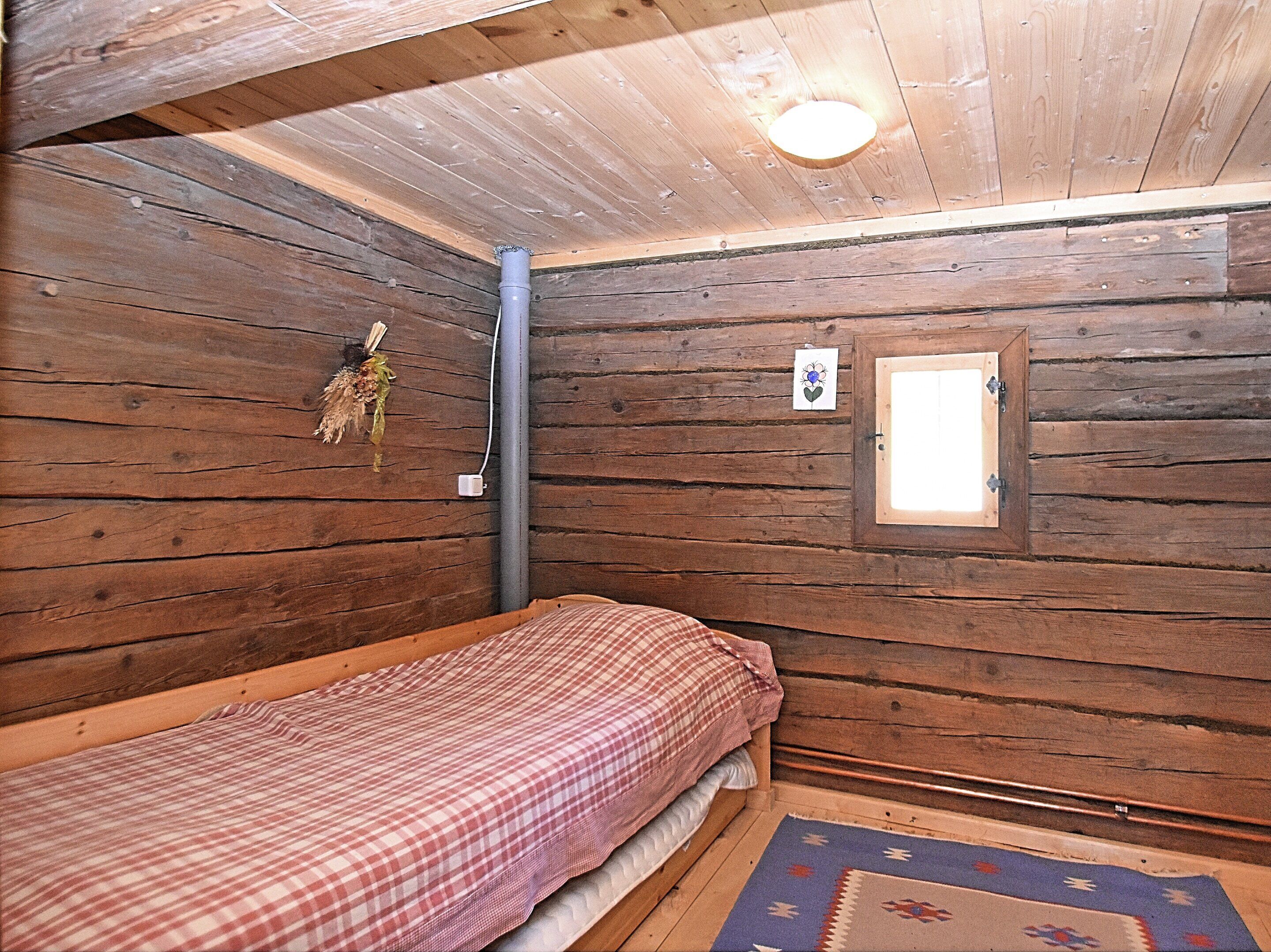 A small, wooden cabin room with a single bed, a window, and a wall-mounted light fixture.