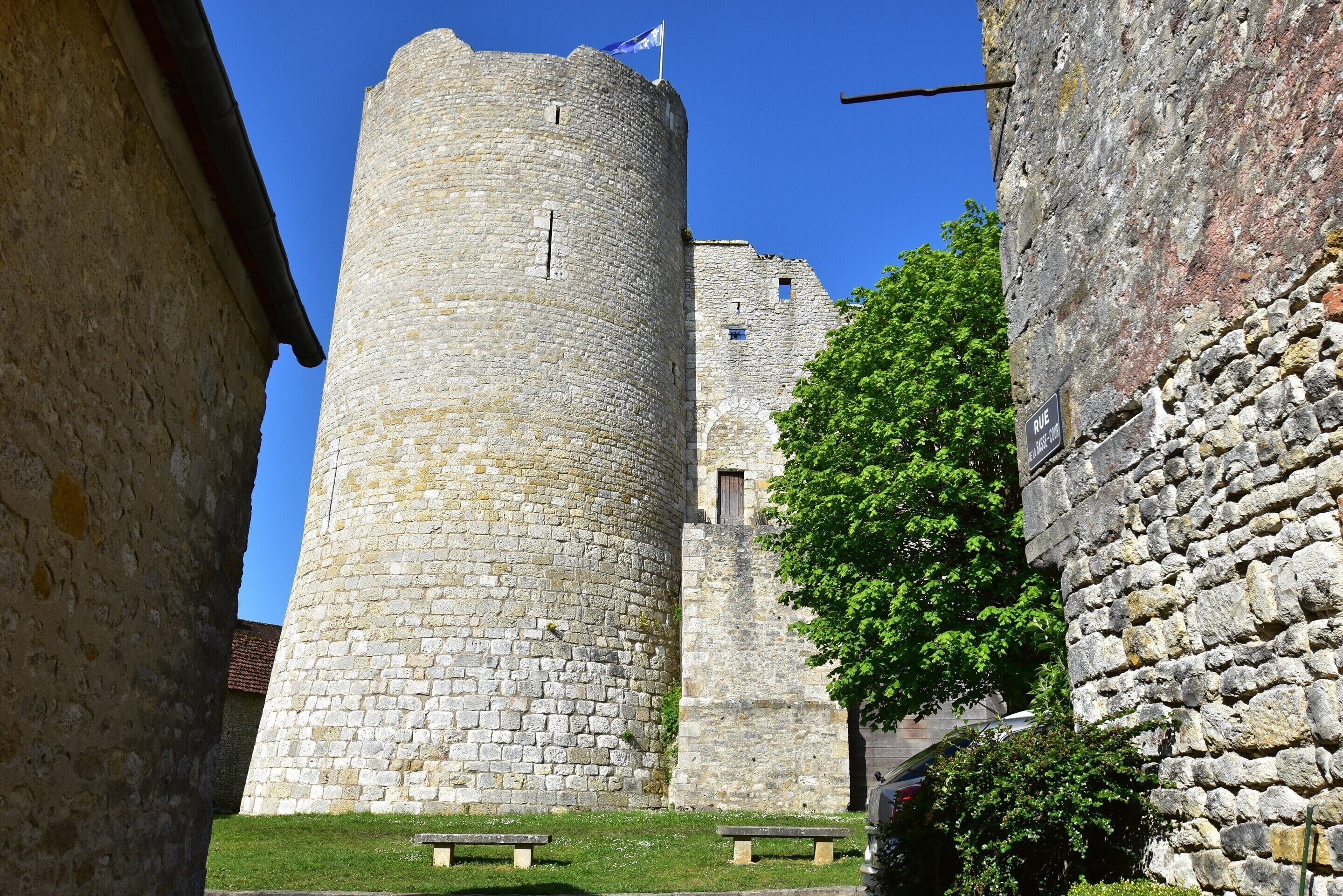 A stone tower with a flag, a tree, and a bench in a courtyard.