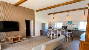 Un salon moderne comprenant un canapé, un téléviseur, un mur recouvert de panneaux en bois, un coin repas, une cuisine et des poutres apparentes en bois.