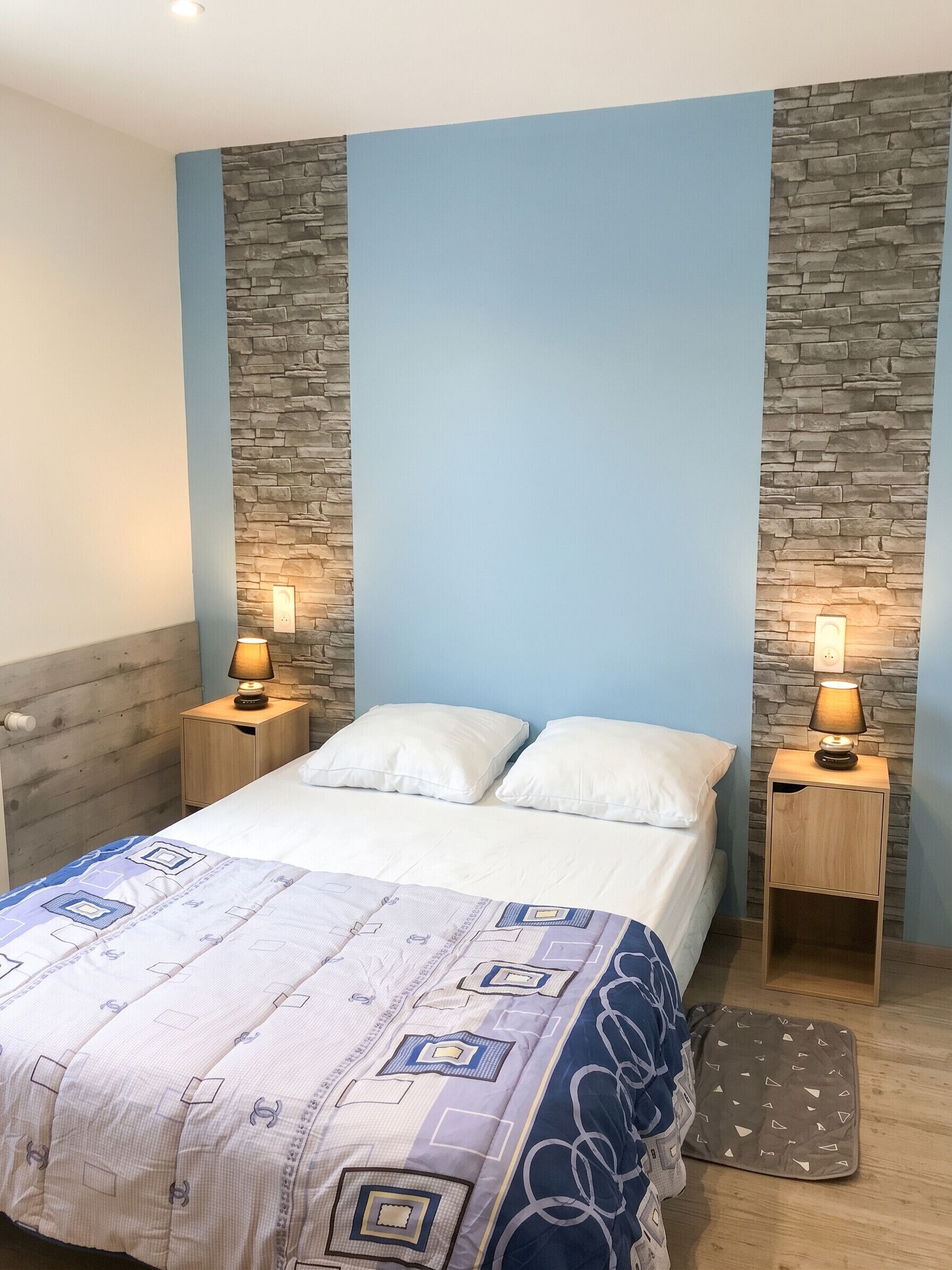 Une chambre à coucher comprenant un lit, deux lampes de chevet, une table de chevet, une tête de lit en bois et un mur d’accent en pierre.
