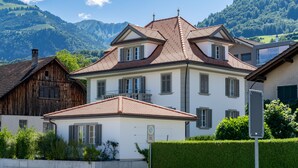 Une maison à deux étages, dotée d’un toit en pente, de nombreuses fenêtres avec volets et d’une cheminée bien visible.