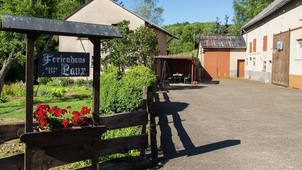 A building with a sign reading "Ferienhaus Lutz" and a wooden bench with red flowers. - Eifel Retreat Near Volcanic Lakes (Ulmen)