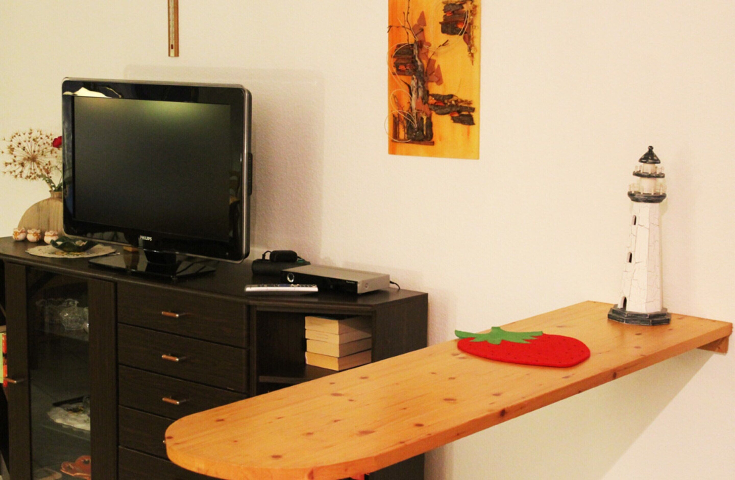 A living room with a TV on a stand, a small table with a red object, and a wall-mounted shelf with a decorative item.