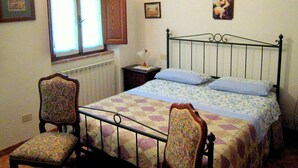 A bedroom with a bed, two chairs, a nightstand, a window, and a painting on the wall. - Tuscan Charm: Frassini Cottage (Chiusdino)