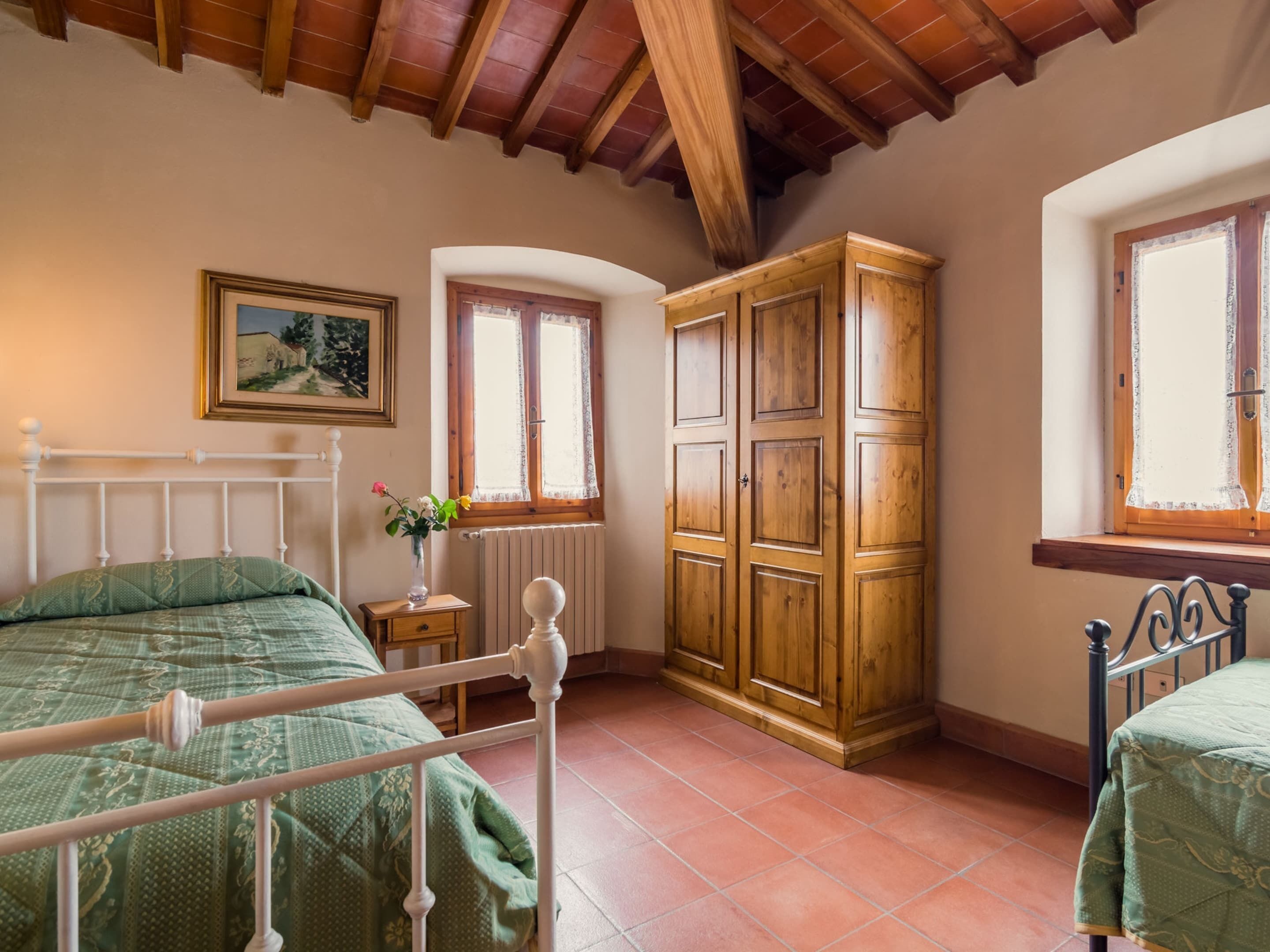 A bedroom with a wooden bed frame, a wooden wardrobe, a radiator, a window with lace curtains, and a framed picture on the wall.