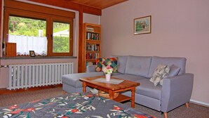 Ein gemütliches Wohnzimmer mit einem grauen Sofa, einem Holztisch, einem gesteppten Teppich, einem Bücherregal und einem Fenster mit Blick auf Grünflächen.