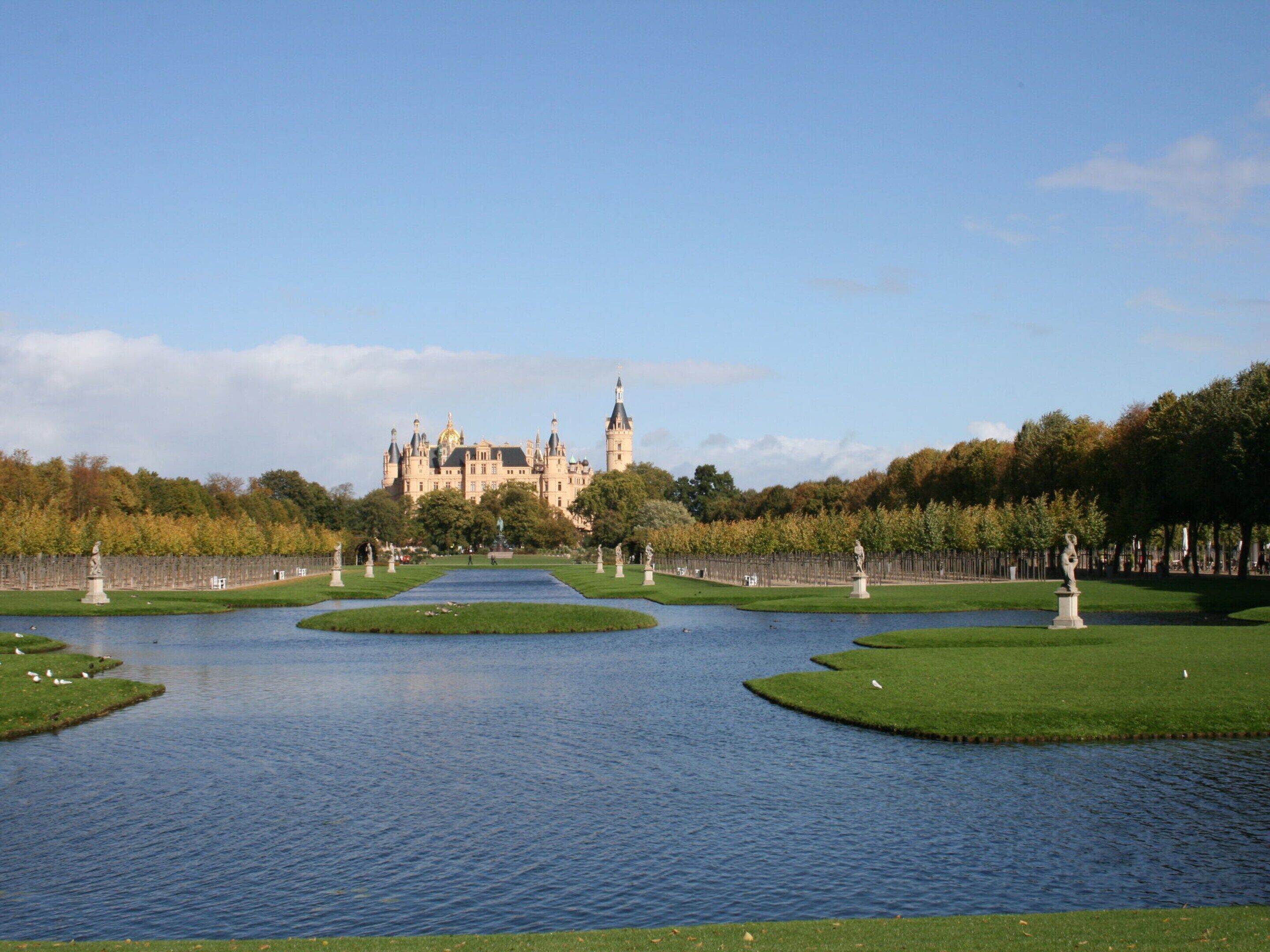 A large, historic building with a symmetrical garden layout, featuring a central water feature and statues.