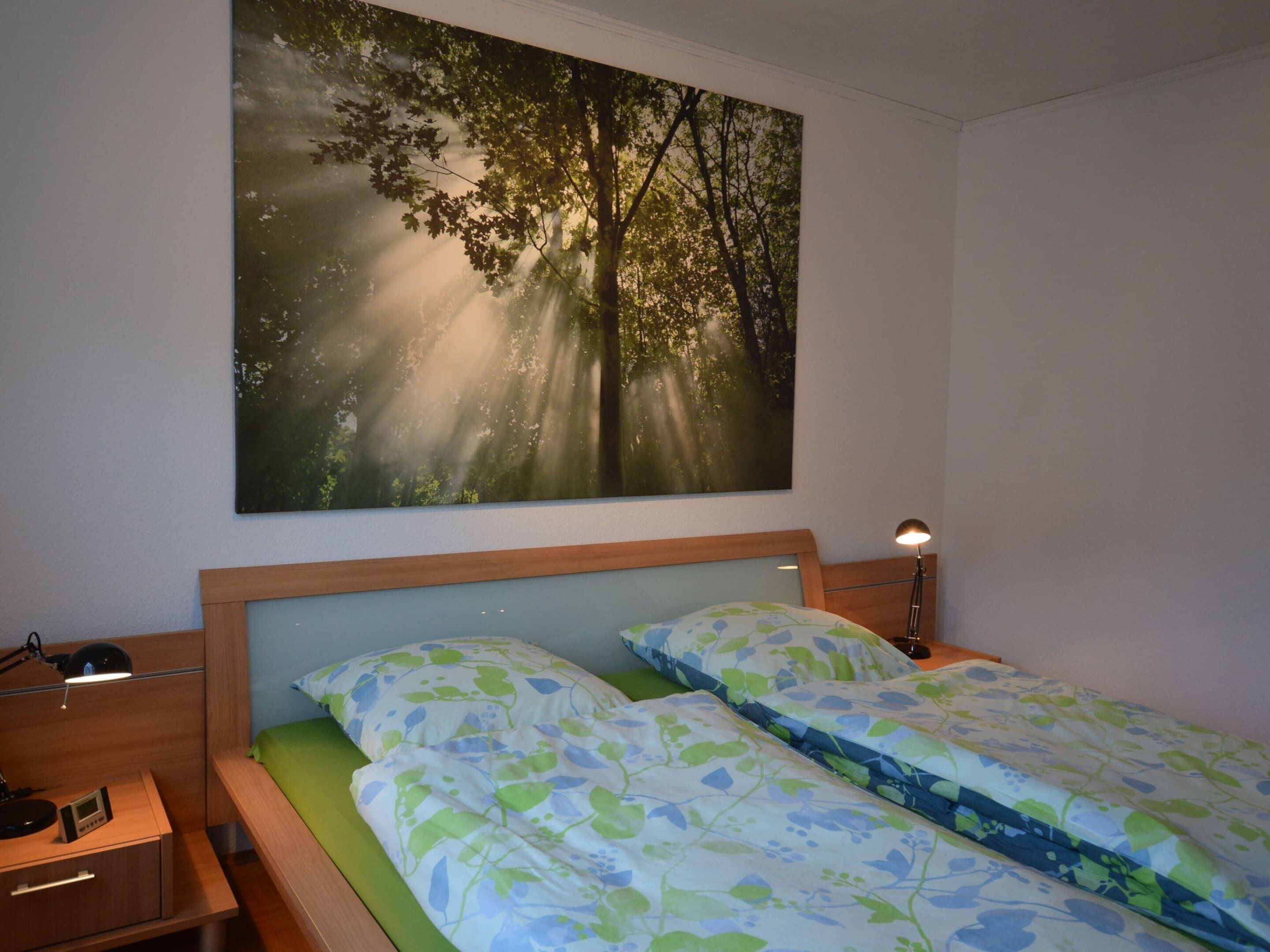 Une chambre à coucher avec un lit, une table de chevet, une lampe et un grand tableau sur le thème de la forêt accroché au mur.