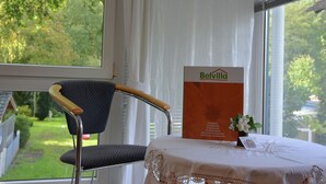 Huone, josta avautuu näkymä vehreään maisemaan. Huoneessa on tuoli, pöytä pitsiliinavaatteella ja kyltti, jossa lukee sana ”Belvilla”.
