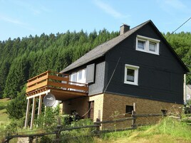 Una casa de dos pisos con fachada oscura, una terraza de madera y una antena parabólica.