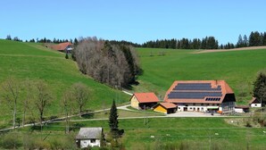 A rural landscape with a large building featuring a solar panel roof, surrounded by green fields and trees. - Ferienhaus, Schwarzwald (Sankt Georgen im Schwarzwald)