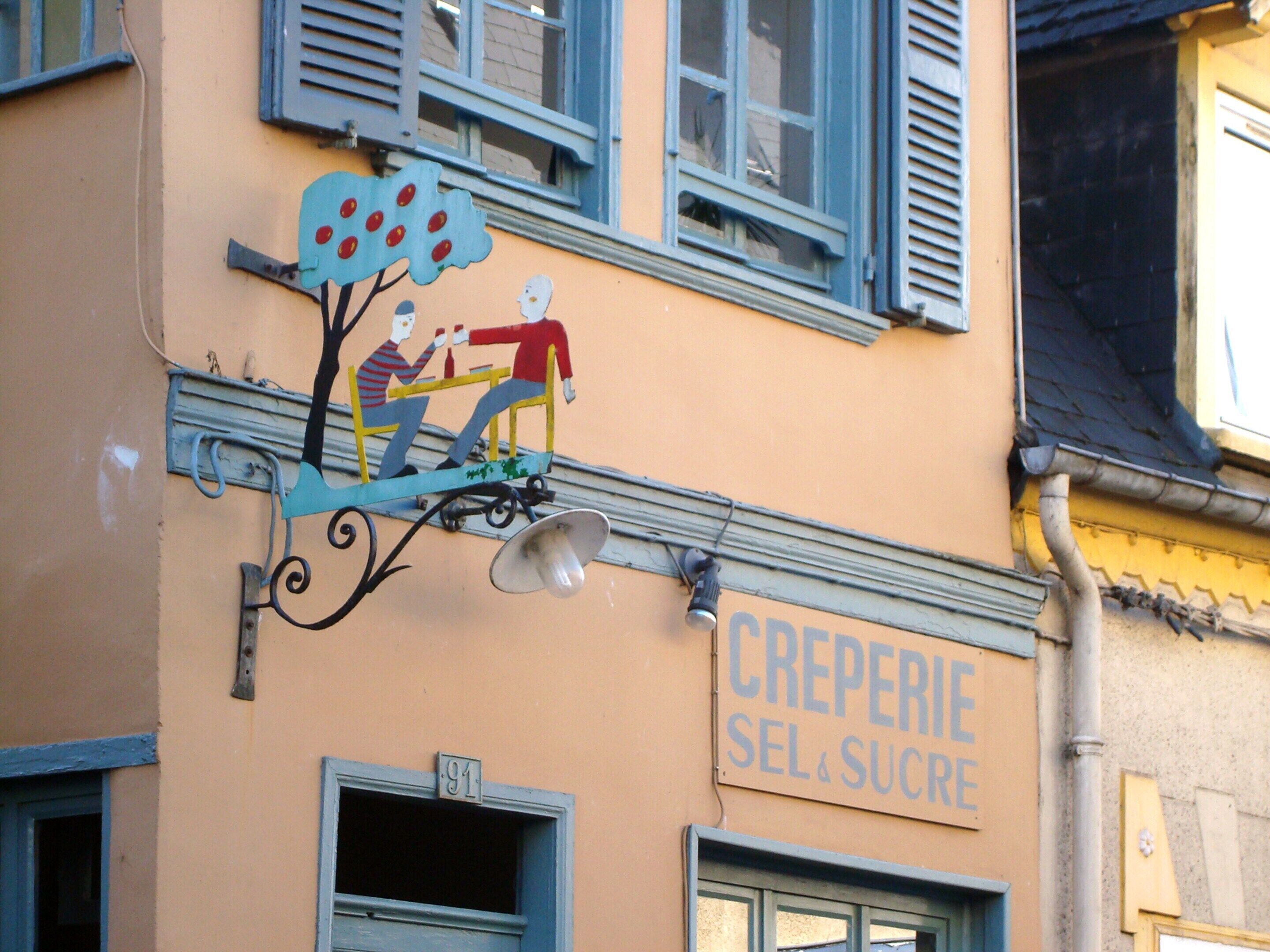 Ein Gebäude mit einem Schild, auf dem 'CREPERIE SEL SUCRE' steht, und einem Wandgemälde von zwei Personen in einem Wagen.