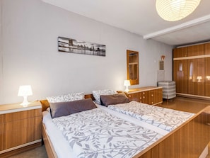 A bedroom with two beds, a wooden wardrobe, a bedside table with a lamp, and a cityscape painting on the wall.