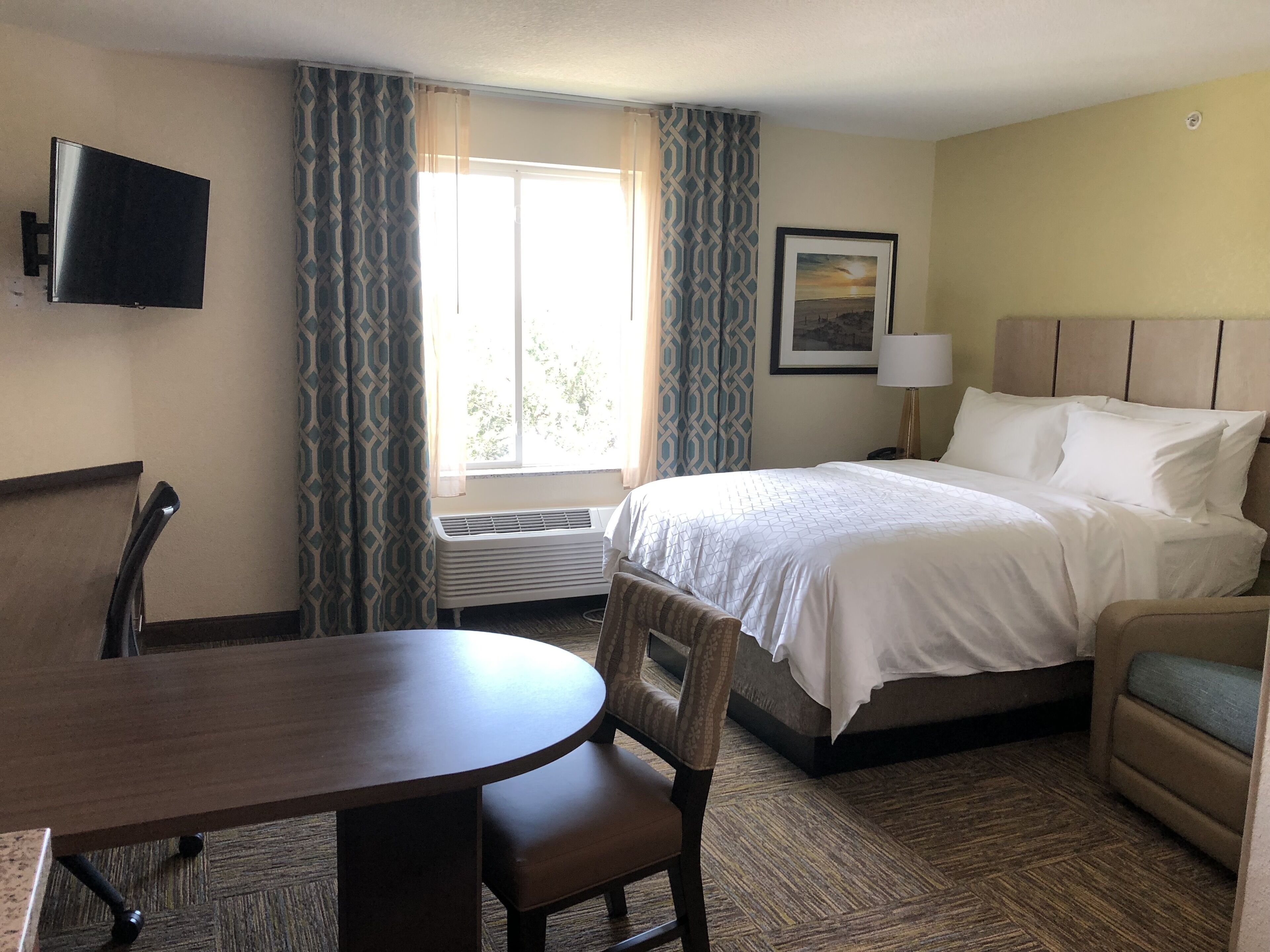 A hotel room with a bed, a desk, a chair, a TV, a window with curtains, and a lamp.