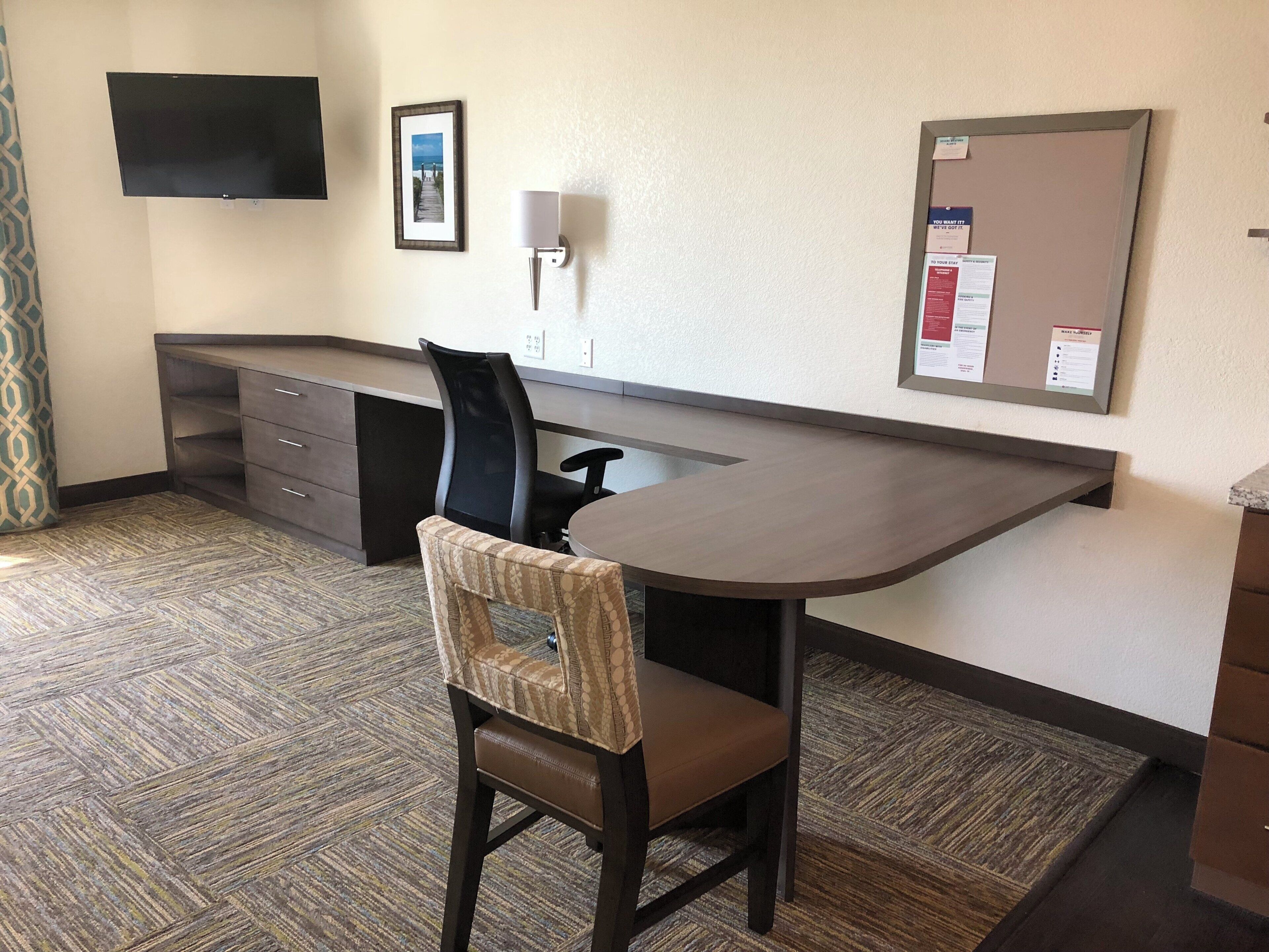 A hotel room with a desk, a chair, a flat-screen TV mounted on the wall, a bulletin board with notices, and a patterned carpet.