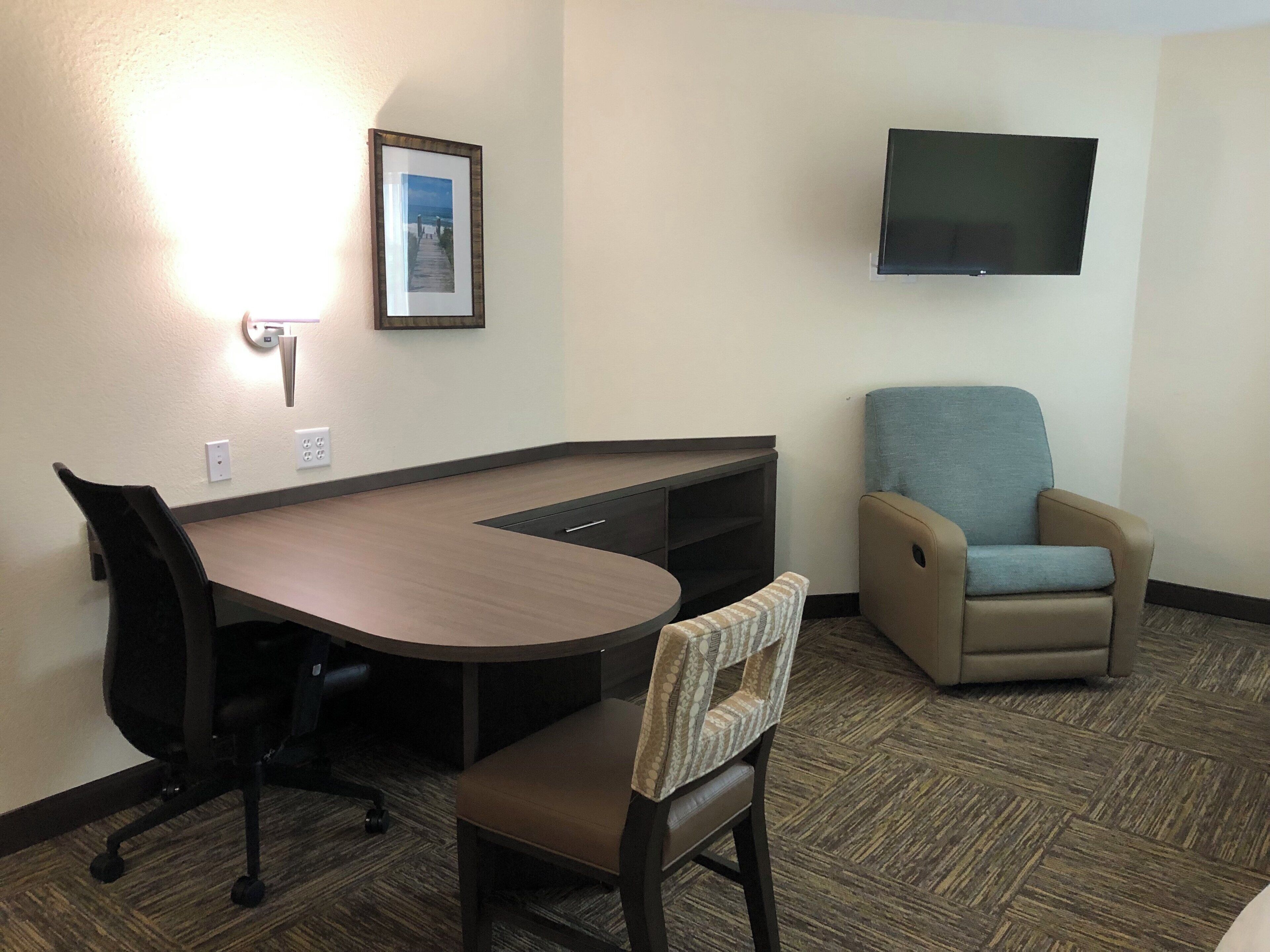 A hotel room with a desk, a chair, a television mounted on the wall, and a framed picture on the wall.