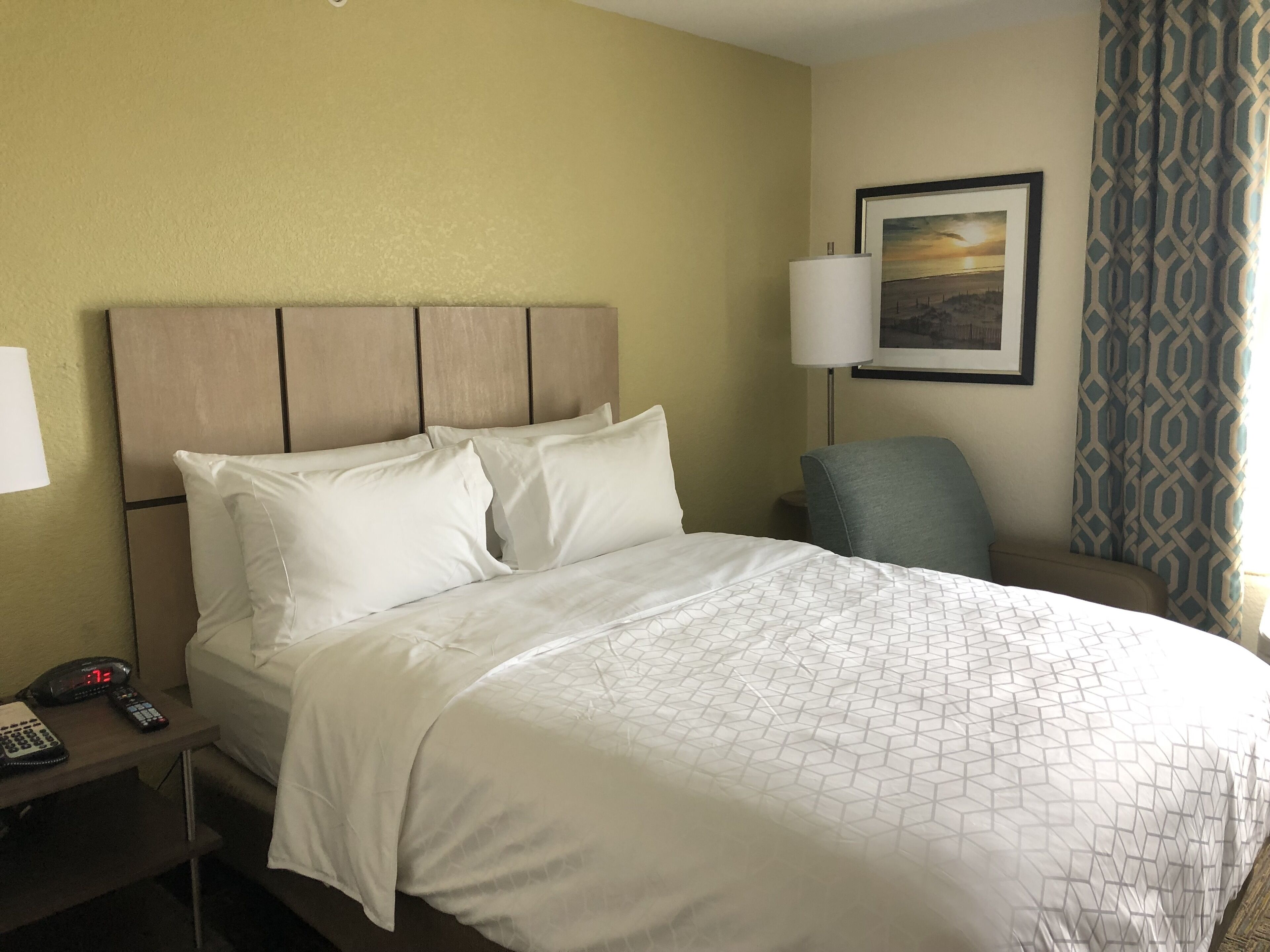 A hotel room with a bed, a nightstand with a phone and remote, a chair, a lamp, and a picture on the wall.