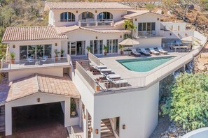 A modern two-story house with a large swimming pool, multiple balconies, and outdoor seating areas.