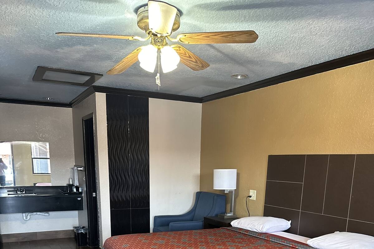 A hotel room with a bed, a desk, a chair, a ceiling fan, and a bathroom with a mirror and sink.