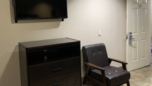 A room with a dark wooden dresser, a leather chair, a mounted television, and a white door with a number plate. - Luxury Inn and Suites (Houston)