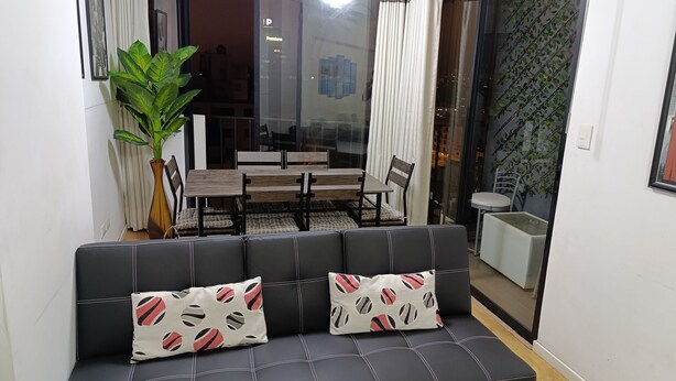 A modern living room with a black sofa, two patterned cushions, a large green plant, and a dining area with a table and chairs. - Pet Friendly Apart(9 Person max)+Pool+Grill+Billiards+Gym (Lince)