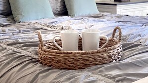 Un lit bien fait, avec un panier tressé contenant deux tasses blanches, une tête de lit et une lampe de chevet.