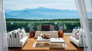 A balcony with a view of mountains and greenery. There are two white sofas with patterned cushions, a wooden coffee table, and two wooden side tables.