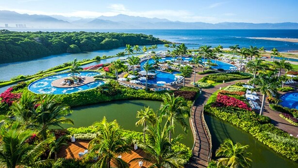 A resort with multiple swimming pools, lush greenery, and a view of the ocean and mountains.