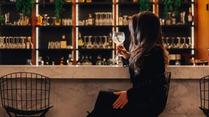 Eine Person sitzt an einer Bar, im Hintergrund Regale mit Flaschen und Gläsern.