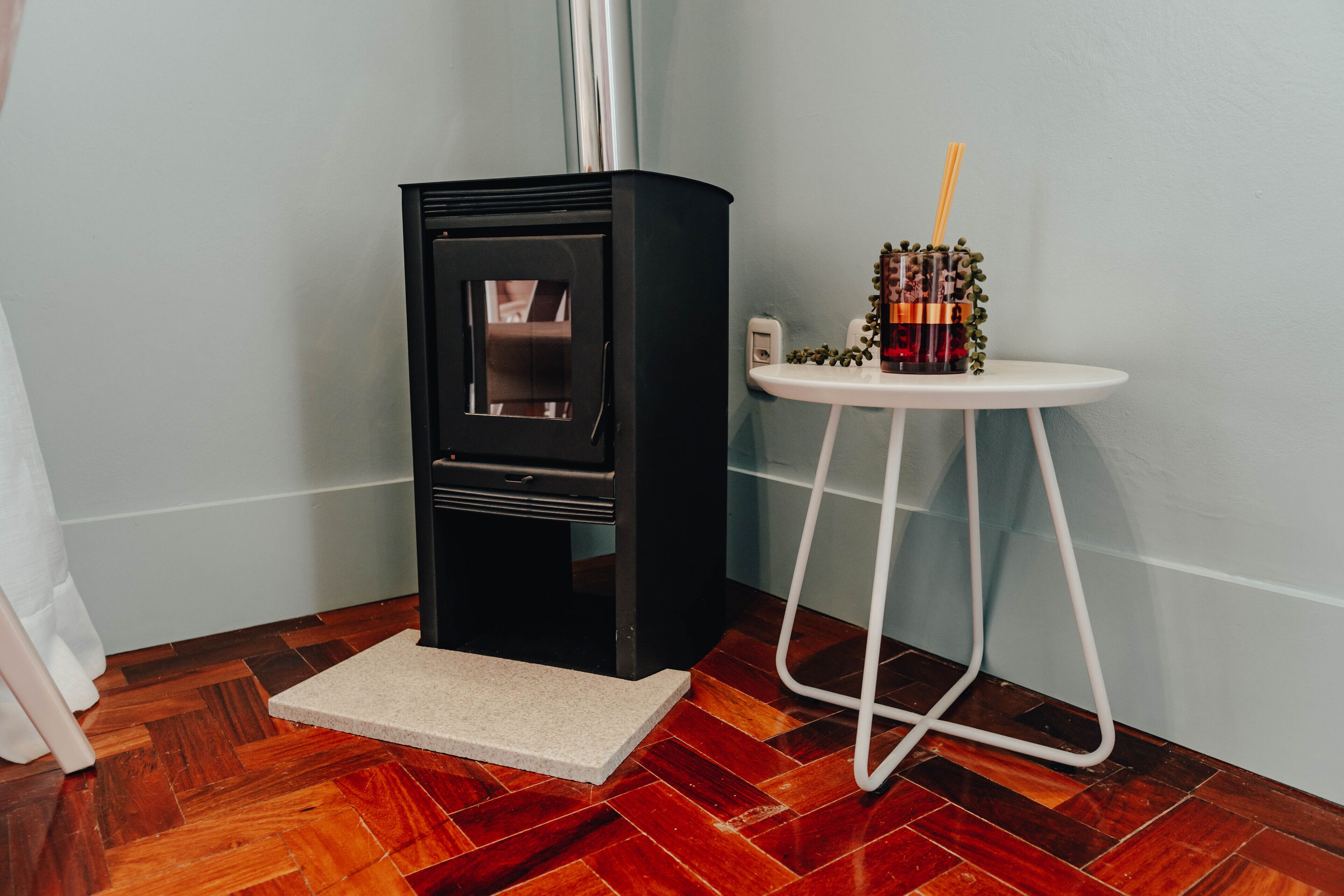 Una habitación de esquina con chimenea de leña, una pequeña mesa blanca con una bebida y suelo de madera.