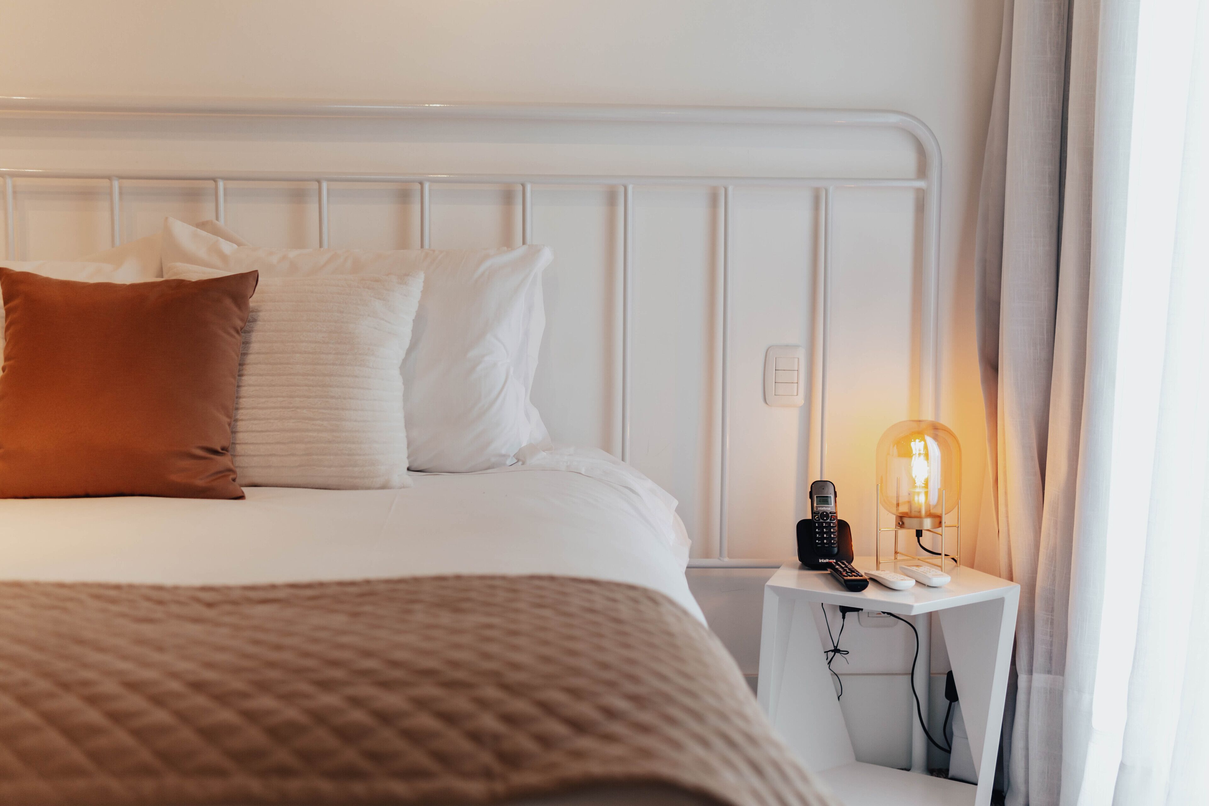 A neatly made bed with white and brown pillows, a bedside table with a lamp, and a power outlet.