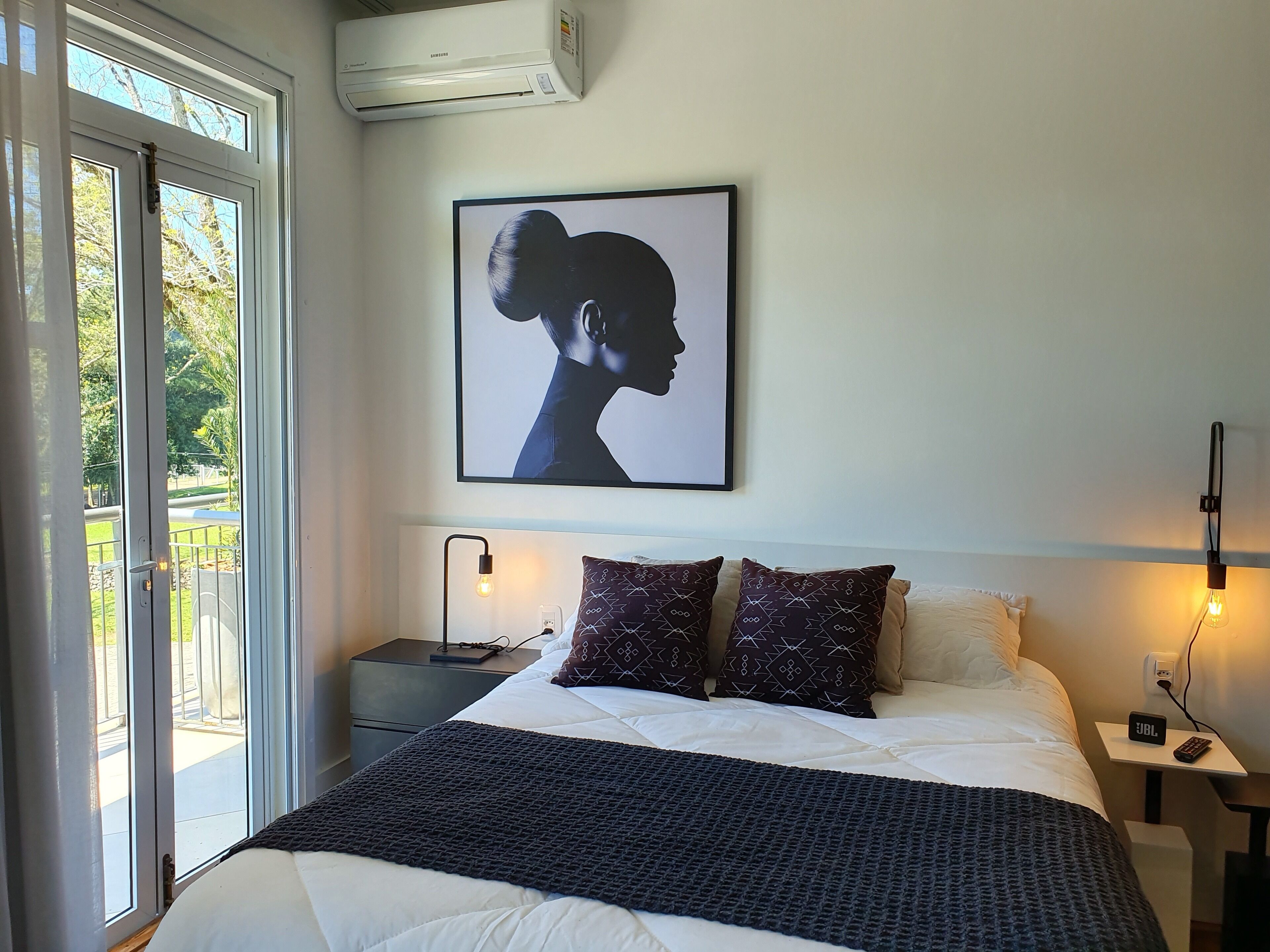 Et soveværelse med seng, puder, et billede på væggen, sengeborde med lamper og en aircondition.