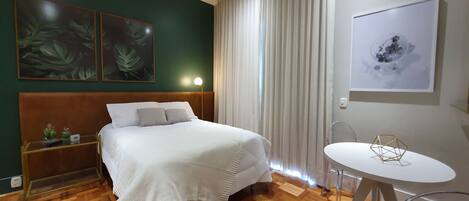 A hotel room with a bed, a bedside table with a lamp, a round table with chairs, and artwork on the walls.