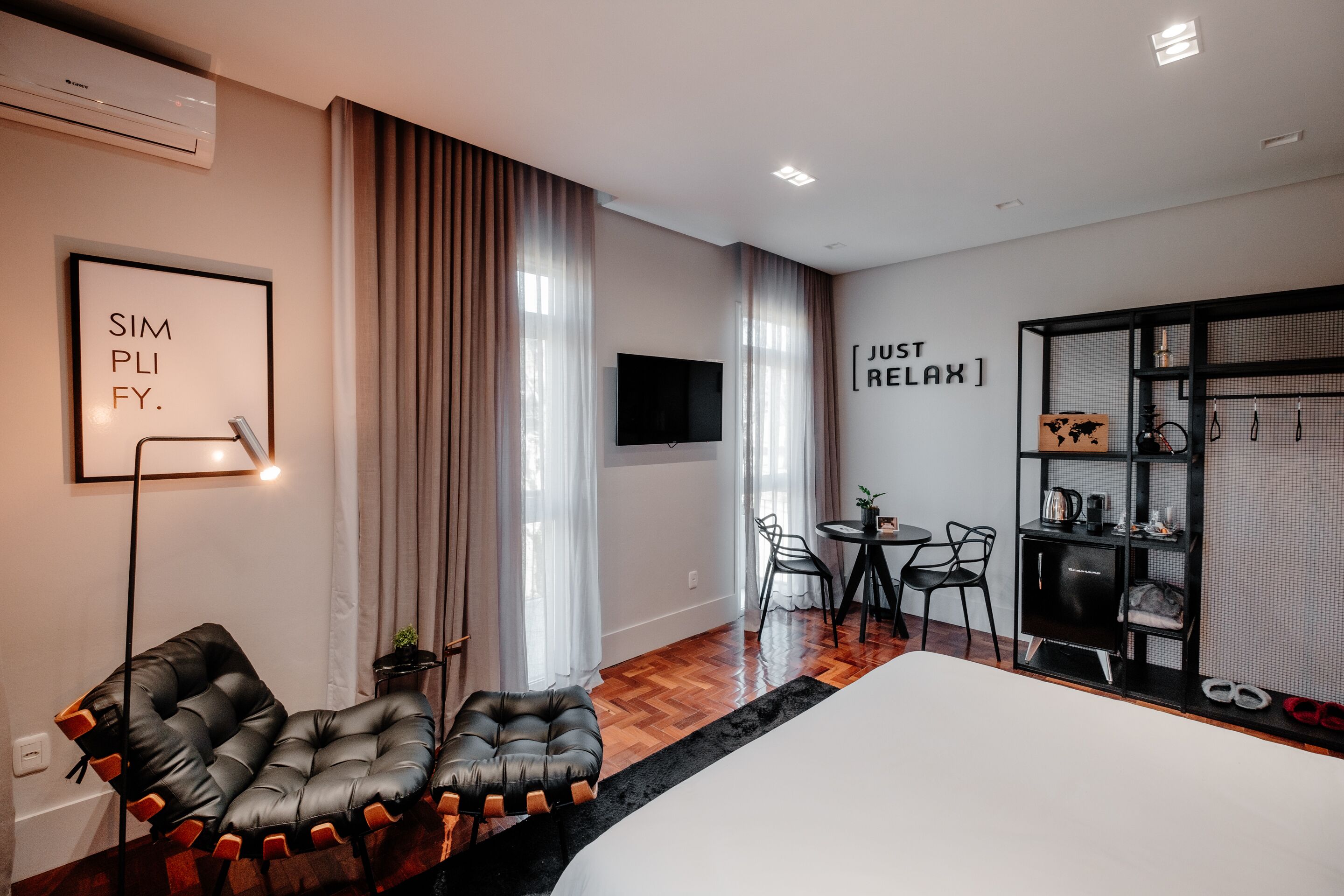 A modern hotel room with a large bed, a black leather chair, a TV, and a small dining area with chairs and a table.