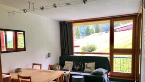 Et værelse med et træbord og stole, en sofa med hynder, et lille rundt bord og udsigt til et grønt landskab gennem et stort vindue.