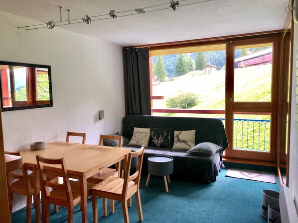 A room with a wooden dining table and chairs, a sofa with cushions, a small round table, and a view of a green landscape through a large window.
