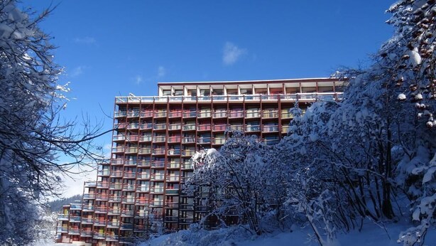 En bygning med flere etager og balkoner, omgivet af sneklædte træer og en klar himmel.