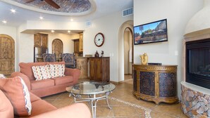 Amplia sala de estar con ventilador de techo, chimenea, televisión y una cocina al fondo.