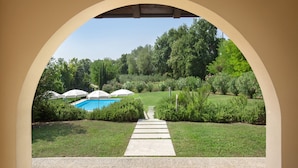 Un percorso conduce a una zona piscina incorniciata da un arco, immersa nel verde e ombrelloni.