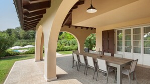 Una veranda esterna con tavolo e sedie in legno, circondata da archi e con vista su una piscina e vegetazione.