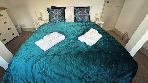 Una cama con una colcha verde azulada, dos toallas blancas dobladas y almohadas estampadas.