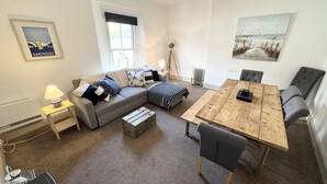 A cozy living room with a grey sofa, a wooden coffee table, a dining table with chairs, a lamp, and a framed picture on the wall.