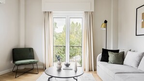Ein modernes Wohnzimmer mit einem runden Tisch, einem grünen Sessel, einem weißen Sofa mit Kissen und Blick auf Grünanlagen durch große Fenster.