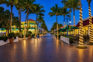 A paved walkway lined with palm trees and outdoor seating areas, leading to a building with a green awning and a sign that reads 'MANGO BAY'.