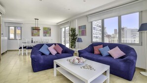 A modern living room with two blue sofas, a white coffee table, and a dining area with a white table and chairs. - Apartment Capri - PlusHolidays (Calpe)