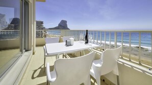 A balcony with a table and chairs, overlooking a beach and a rocky outcrop. - Apartment Capri - PlusHolidays (Calpe)