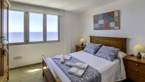 A bedroom with a bed, two bedside tables, a nightstand, a window with a view of the sea, and a ceiling light. - Apartment Capri - PlusHolidays (Calpe)