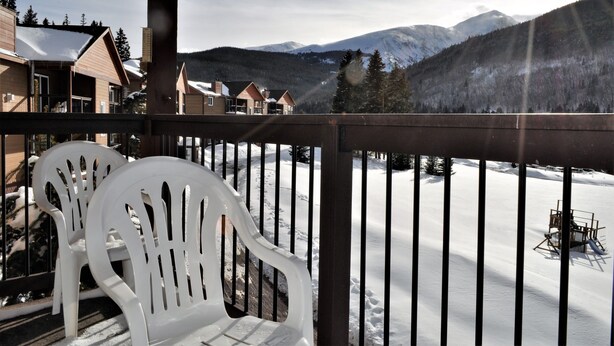 Une terrasse en bois avec des chaises en plastique blanches, donnant sur un paysage enneigé, avec des montagnes et des arbres.