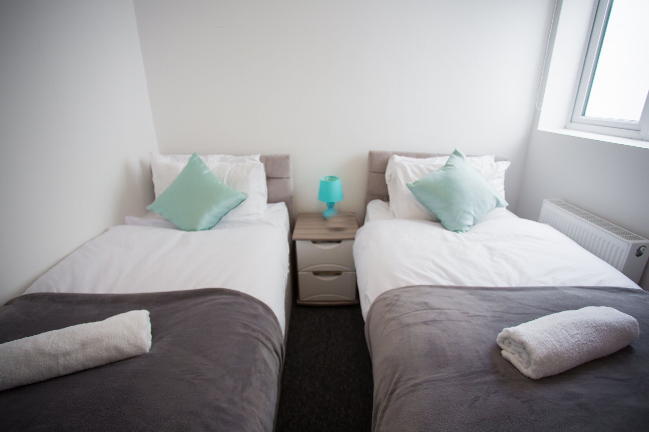 Habitación doble con dos camas, cada una con una almohada y una toalla. Hay una mesita de noche con una lámpara entre las camas.
