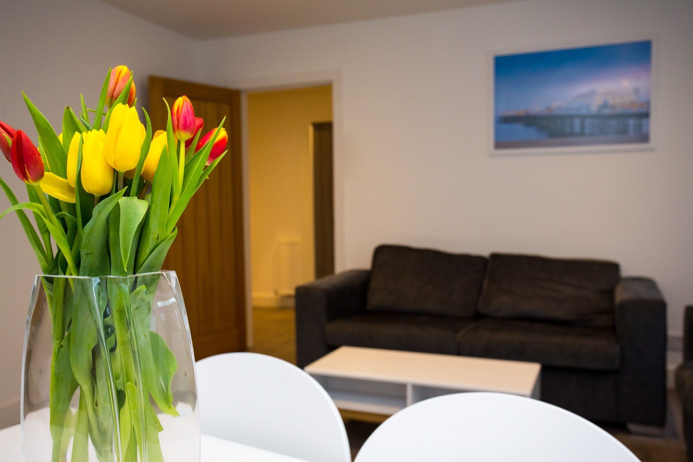Un salón con un jarrón de tulipanes sobre una mesa blanca, un sofá marrón oscuro y un cuadro en la pared.